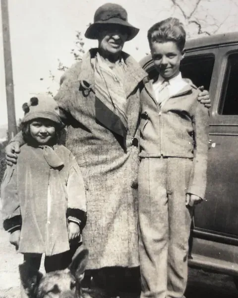 An old black-and-white photo of a woman in a hat with two children beside a car.